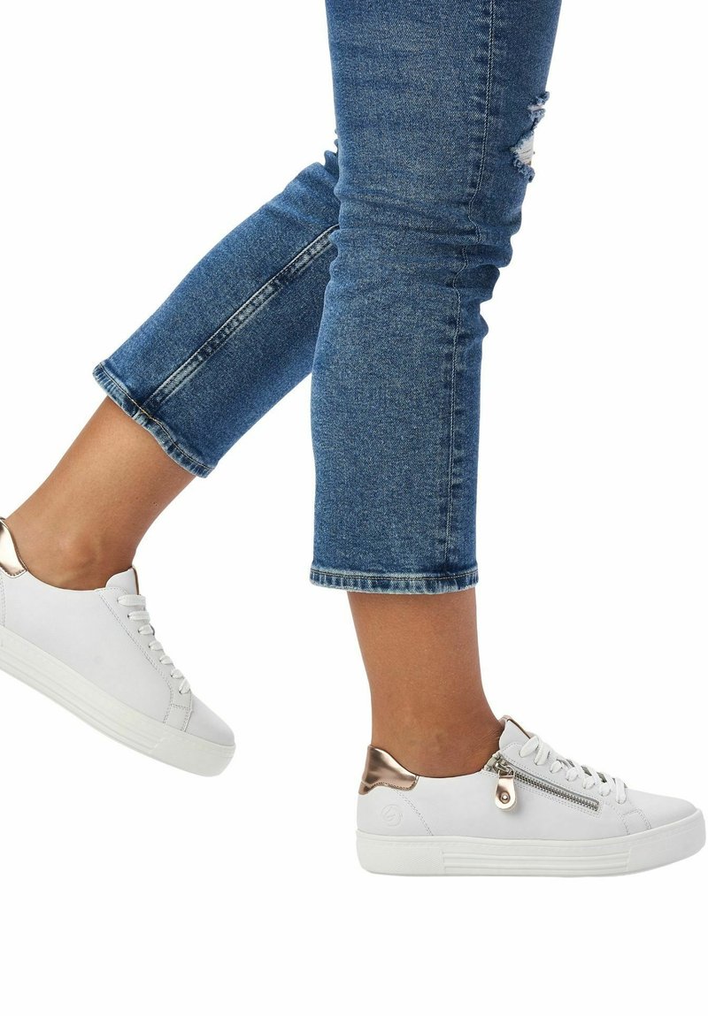 Baskets blanches avec un accent en rose gold brillant au talon et une fermeture éclair sur le côté, dotées d'une semelle texturée et de lacets. Portées avec un jean bleu retroussé.