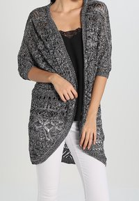 Grå stickad cardigan med öppen front, som har en lös, luftig design och intrikata spetsmönster. Bärs över en svart topp och vita byxor.