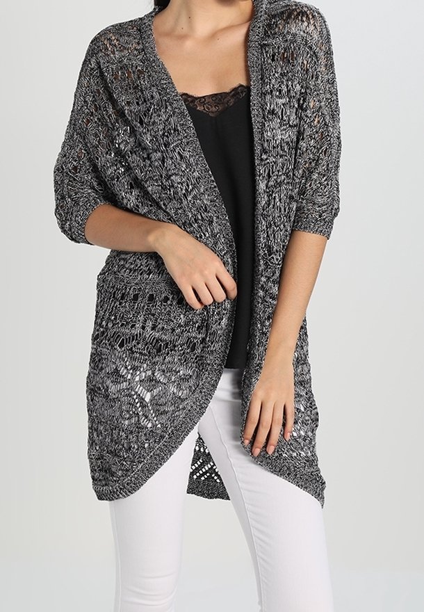 Grå stickad cardigan med öppen front, som har en lös, luftig design och intrikata spetsmönster. Bärs över en svart topp och vita byxor.