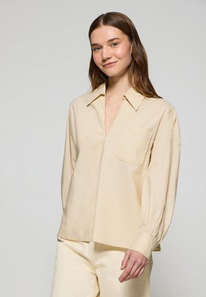 Camicia beige a maniche lunghe con scollatura a V, polsini con bottone e taschino sul lato sinistro del petto. Tessuto morbido con una consistenza liscia e una vestibilità comoda.
