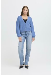 Cardigan blu lavorato a maglia con scollo a V, chiusura con bottoni e polsini a coste, abbinato a jeans azzurri e scarpe nere con tacco.