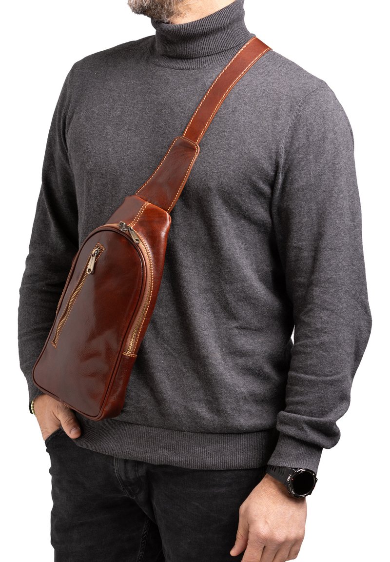 Borsa a tracolla in pelle marrone con finitura liscia, tasca frontale con zip e cuciture a contrasto. Indossata a tracolla sopra un maglione grigio.