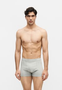 Jeune homme aux cheveux courts et foncés portant un boxer Calvin Klein gris clair, debout devant un fond blanc uni, face à l'avant.