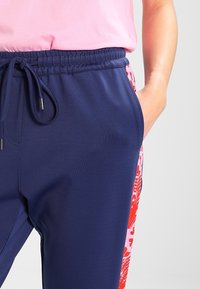 Mörkblå joggers med en strukturerad yta, prydd med ett färgglatt blommönster i röd och rosa på sidan. Elastisk midja med dragsko.