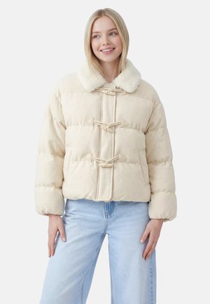 Ung kvinde iført en cremefarvet dunjakke med lukninger med snøre og en kunstpelskrave, kombineret med lyseblå vidde jeans.