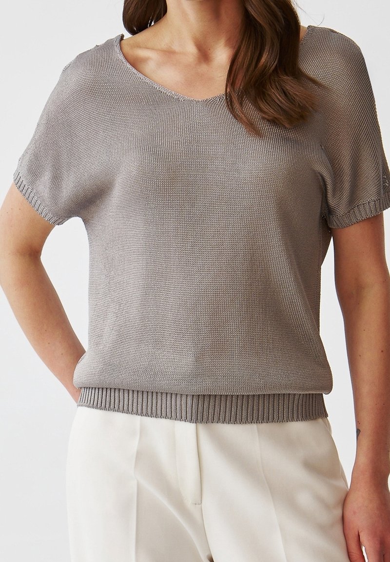 Femme portant un haut gris en maille à manches courtes avec col en V et un pantalon blanc taille haute, debout avec une main dans la poche.