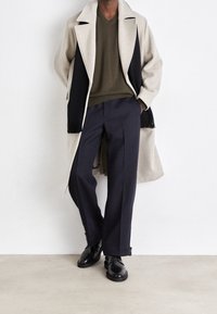 Manteau en mélange de laine beige avec accents noirs, pull en maille vert olive, pantalon large bleu marine, et chaussures en cuir noir. Détails à franges sur le manteau.