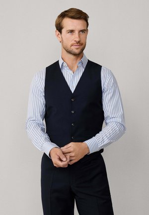 Hombre con chaleco azul marino sobre camisa de rayas blancas y azul claro, ajustándose las manos frente a fondo neutro.