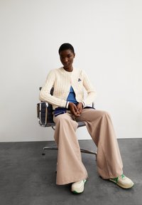 Cremfarbener, strukturierter Strickcardigan mit rundem Ausschnitt und navyfarbenem Logo. Kombiniert mit einem blauen Oberteil und hellbeigen, weiten Hosen, ergänzt durch Sneakers.