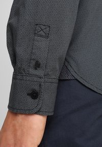 Donkergrijze shirtmouw met een gepunkteerde textuur, knoopsluitingen op de manchetten en accentstiksels. De stof is glad en heeft een gestructureerde pasvorm.