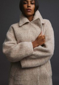 Béžový vlněný kabát s texturovaným povrchem, velkým límcem a nadměrnými rukávy, zdobený jemnými barevnými skvrnami a volným střihem.