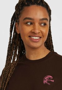 Sweatshirt marron avec un col rond, des poignets côtelés et un logo rose sur la poitrine. Fabriqué en tissu doux avec une texture lisse.