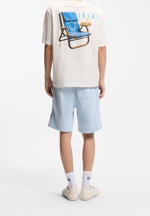 Personne portant un t-shirt blanc avec un imprimé de chaise bleue et boisson, short bleu clair, chaussettes blanches et baskets blanches, debout de dos.