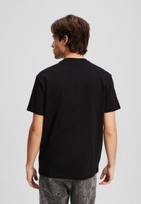 Melna īsa piedurkņu T-krekls, kas izgatavots no kokvilnas. Tam ir ribota apaļa kakla izgriezums un brīvs piegriezums. Aizmugurējā skatījumā redzams gluds tekstūrējums, bez rakstiem vai akcentiem.