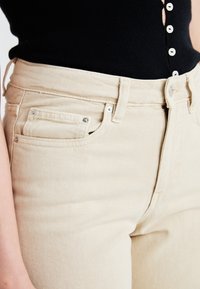 Beiga jeans med hög midja, framficka, metalldetaljer och en strukturerad yta. Stylad med en svart topp.