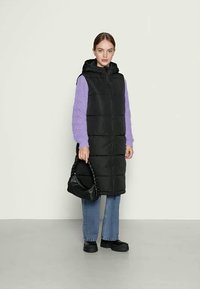 Mladá žena v dlhom čiernom pufferveste, fialovom svetri z pleteniny, modrých džínsach, čiernych platformových topánkach, s čiernou pletenou kabelkou.