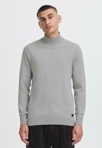 Maglione grigio a collo alto realizzato in tessuto a maglia fine, con maniche lunghe, polsini e orlo a costine, e una piccola etichetta con logo in fondo.