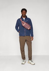 Navy fleece zip jacket, tan pants, and beige sneakers. Pink sling bag with "Patagonia" branding. Standing against a neutral background.