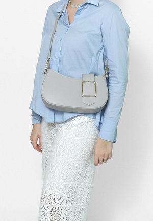 Femme portant une chemise bleue claire boutonnée et une jupe blanche à motifs, portant un sac à bandoulière gris avec une grande boucle dorée.