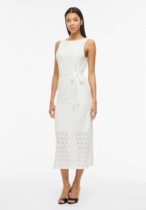 Kobieta w białej, bezrękawnikowej dzianinowej sukience midi z paskiem wiązanym na boku i czarnych sandałach na obcasie, stojąca na jednolitym białym tle.