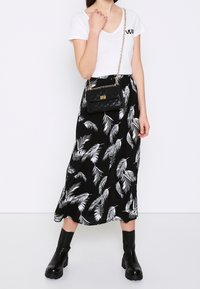 Nainen, jolla on päällään valkoinen lyhythihainen paita, musta hame, jossa on valkoisia lehtikuvioita, mustat nilkkurit ja joka kantaa mustaa tikattua olkalaukkua.