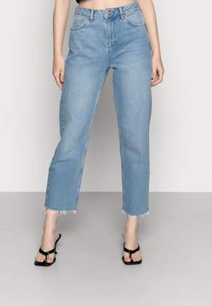 Kobieta ubrana w jasnoniebieskie krótkie jeansy z wysokim stanem i surowymi krawędziami oraz czarne sandały na obcasie z odkrytymi palcami, stojąca na jednolitym tle.