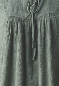 Grünes, strukturiertes Kleid mit einem gerafften Oberteil, das über einen bindbaren Ausschnitt sowie kleine Erhebungen im Stoff für zusätzliche Details verfügt.