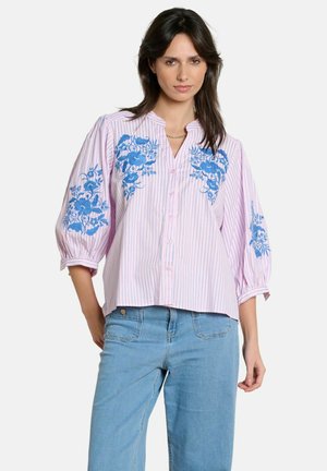 Naine kannab heledat roosat nööpidega pluusi, millel on sinine lilleline tikand ja vertikaalsed triibud, koos helesiniste teksadega.