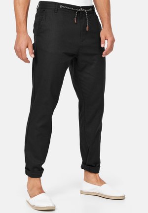 Hombre con pantalones negros remangados con cinturón trenzado y zapatos blancos sin cordones, de pie contra un fondo liso.