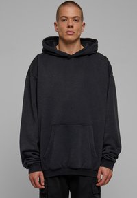 Hoodie nero oversize realizzato in tessuto morbido, con tasca frontale, cappuccio con cordino e polsini a costine, indossato con pantaloni neri larghi.