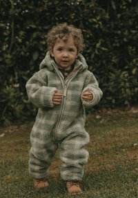 Ljusgrön fleece overall med dragkedja fram, huva och texturerad tyg. Liten person som bär bruna snörkängor, står i gräset.