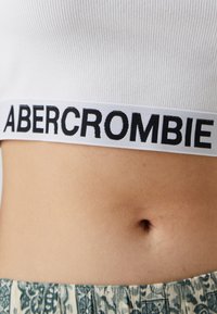 Biely rebrovaný crop top s čiernym logom "ABERCROMBIE" na širokom páse; kombinovaný so vzorovaným, ľahkým spodkom.