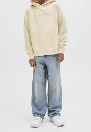 Person iført en cremefarvet hættetrøje med frontlomme, lyseblå vide jeans og hvide sneakers, stående foran en ensfarvet hvid baggrund.