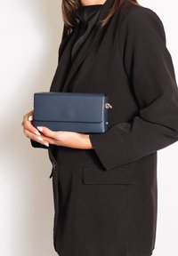 Borsa a mano blu navy con superficie liscia, forma rettangolare e chiusura magnetica. Tenuta in mano accanto a un blazer nero.