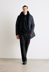 Black puffer jacket worn over a black polo shirt, paired with black trousers and shoes. Model stands against a light background.