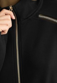 Czarna kurtka na zamek z wysokim kołnierzem, z zamkiem w złotym odcieniu i siateczkowymi wstawkami na ramionach. Gładka tekstura materiału.
