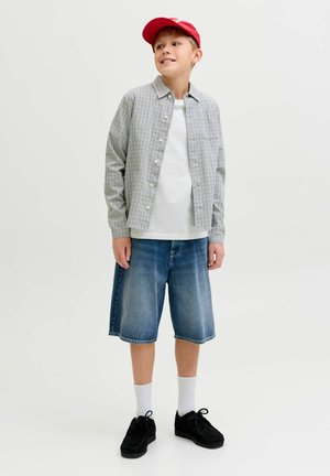 Ung dreng står op, iført rød kasket, ternet skjorte med knapper over hvid t-shirt, denimbukser, hvide sokker og sorte sko, og kigger mod højre.