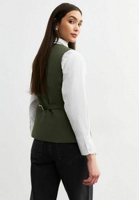 Chaleco ajustado verde oliva con un diseño elegante, que presenta un cinturón en la cintura y una abertura en la parte posterior, combinado con una camisa blanca de manga larga y pantalones negros.