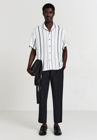 Uomo che indossa una camicia bianca a maniche corte con righe verticali nere, pantaloni neri cropped, mocassini neri, mentre tiene una borsa di pelle nera.