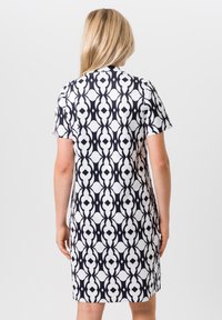 Vrouw met blond haar in een zwart-wit geometrisch patroon jurk met korte mouwen, met de rug naar de camera tegen een witte achtergrond.