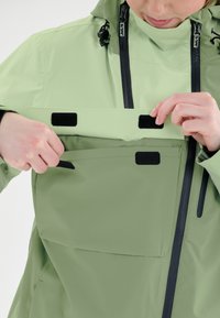 Vaaleanvihreä vedenpitävä takki, jossa on vetoketju ja tarrakiinnityksellä varustetut taskut, säädettävä huppu ja tyylikäs, sileä pinta. Kestäviä suunnitteluseikkoja näkyvissä.