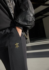 Čierne športové nohavice s bočnými vreckami, s kultivovaným zlatým kvetinovým logom na ľavom stehne, v kombinácii so štruktúrovanou čiernou bomber bundou.