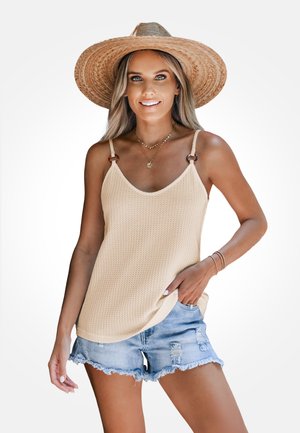 Beiger Waffelstrick-Top mit verstellbaren Trägern und holzähnlichen Akzenten; kombiniert mit abgenutzten Jeansshorts. Neutrale Farbpalette.