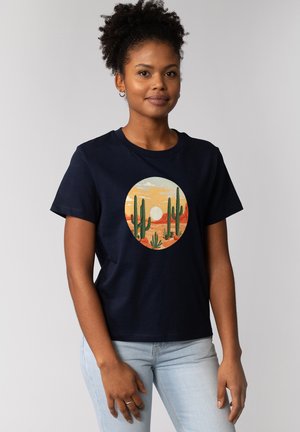 T-shirt en coton bleu marine avec un motif circulaire de paysage désertique comprenant des cactus, des montagnes, un soleil couchant et des couleurs pastel.