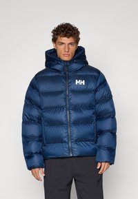 Veste matelassée bleu marine avec des sections quiltées horizontales, un col haut et une fermeture éclair à l'avant. Comprend un logo sur la poitrine gauche.