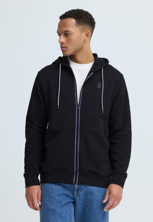 Jonge man met een zwarte hoodie met ritssluiting en capuchon, en lichte blauwe spijkerbroek, kijkend naar links tegen een effen achtergrond.