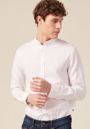 Weißes Leinenhemd mit Stehkragen, langen Ärmeln und durchgehender Knopfleiste, kombiniert mit dunkelblauen Jeans. Schlichtes und minimalistisches Design.