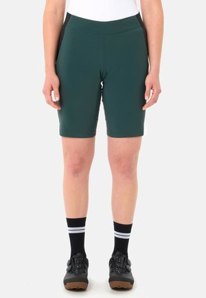 Person trägt dunkelgrüne Radsportshorts, schwarze Socken mit weißen Streifen und schwarze Radsportschuhe und steht vor einem weißen Hintergrund.