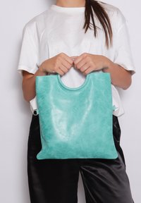 Turquoise leren tote bag met een ronde uitsnijding in het handvat, een gladde textuur en een minimalistisch ontwerp, vastgehouden door een persoon in casual kleding.