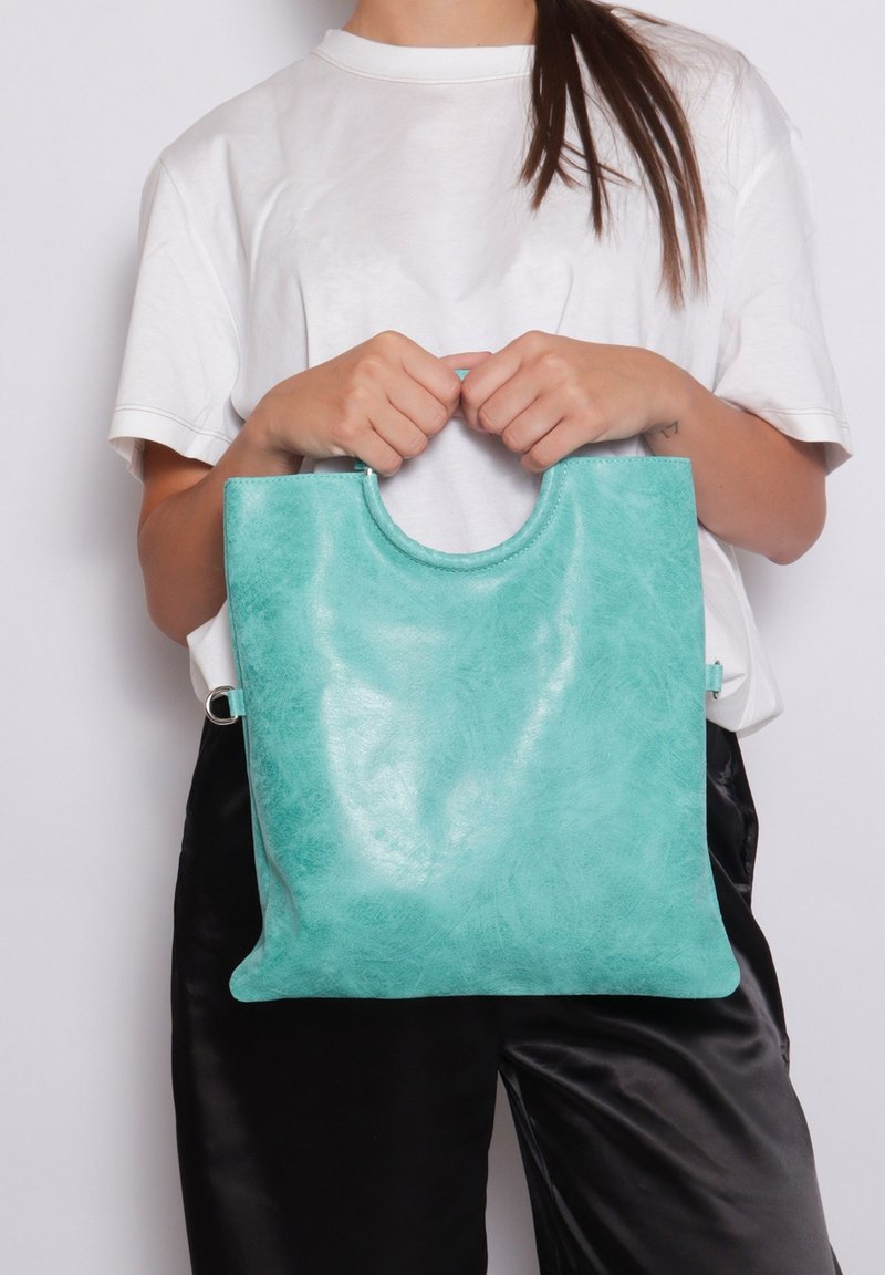 Turquoise leren tote bag met een ronde uitsnijding in het handvat, een gladde textuur en een minimalistisch ontwerp, vastgehouden door een persoon in casual kleding.